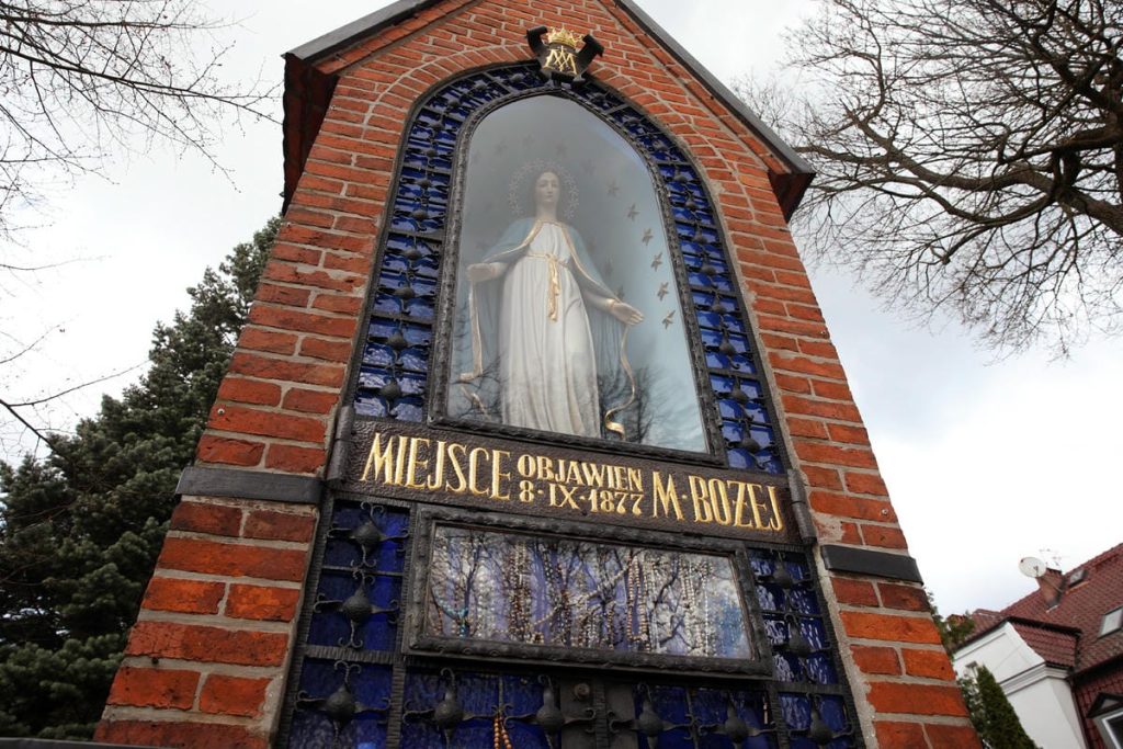 Zabytkowa kaplica w Gietrzwałdzie z obrazem Matki Boskiej i tablicą informacyjną o objawieniach.