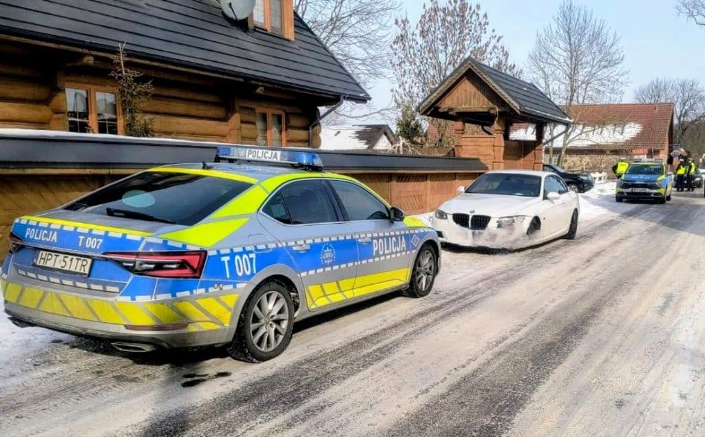 Policja zatrzymuje samochody na zaśnieżonej drodze obok zamarzniętego jeziora, w tle domy i patrol.