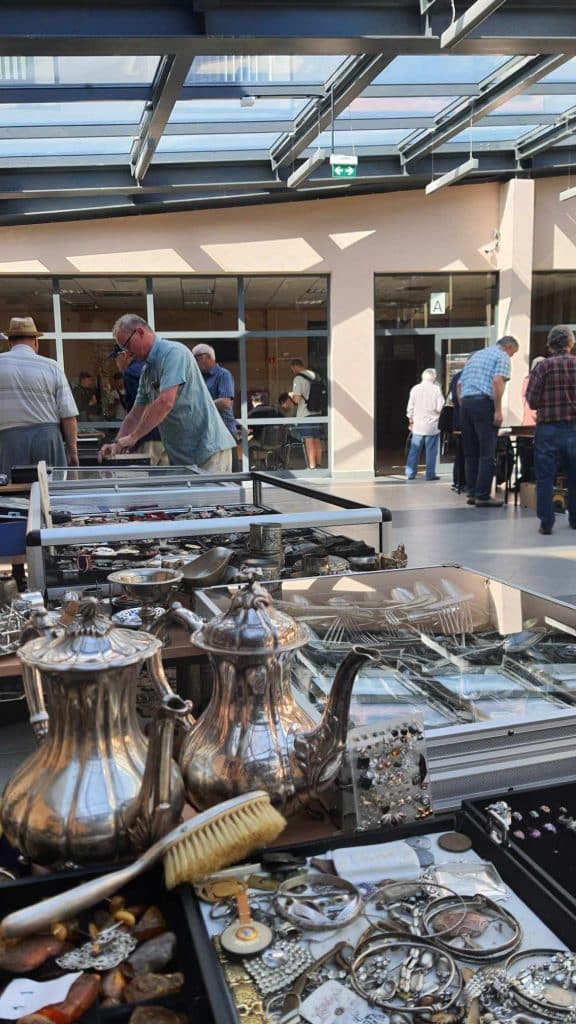 Stoiska z kolekcjonerskimi przedmiotami na giełdzie w Olsztynie, ludzie przeglądają pamiątki w hali.