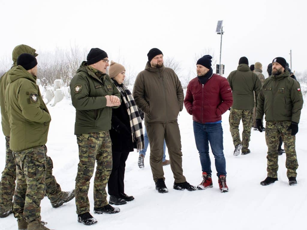 Grupa funkcjonariuszy i przedstawicieli służb na śnieżnym terenie, rozmawiający podczas wizyty na granicy.
