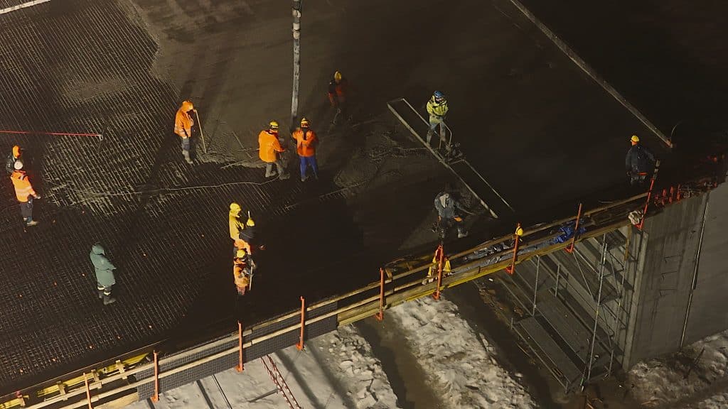 Prace budowlane na moście z ciężkim sprzętem i robotnikami w nocy.