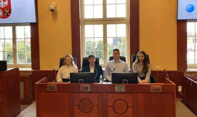 Młodzi ludzie siedzą przy biurku w sali konferencyjnej z dużymi oknami i ekranami na ścianach.