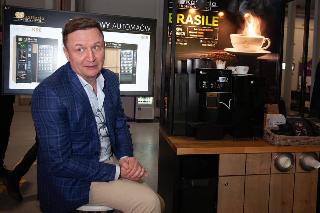 Mężczyzna siedzi przy automacie do kawy na targach w Gdańsku, w tle wyświetlacze i stoisko z kawą.