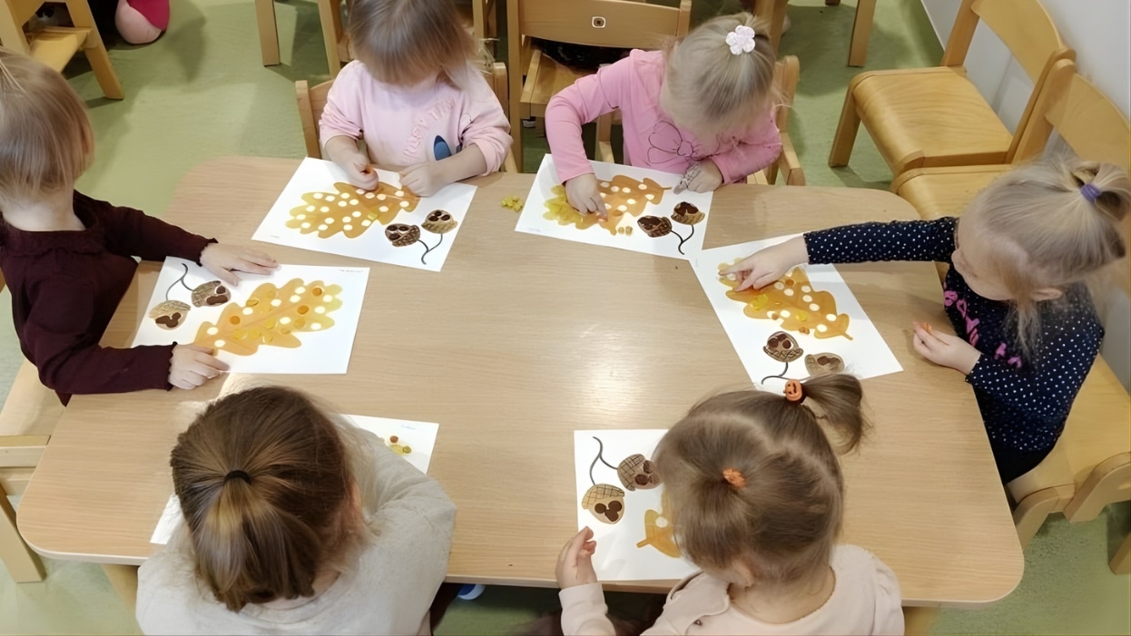 Grupa małych dzieci podczas zajęć plastycznych w żłobku, siedzą przy stole i tworzą prace z papieru i naklejek.