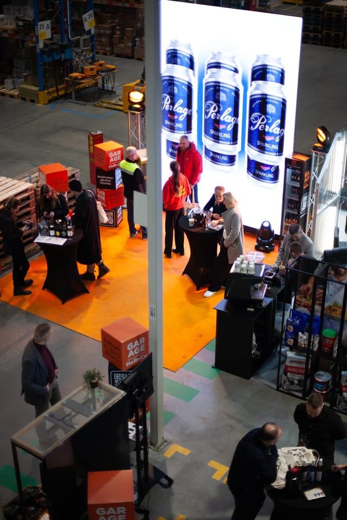 Stoisko z dużym ekranem wyświetlającym reklamy napoj&oacute;w, w tle ludzie rozmawiają i degustują produkty.