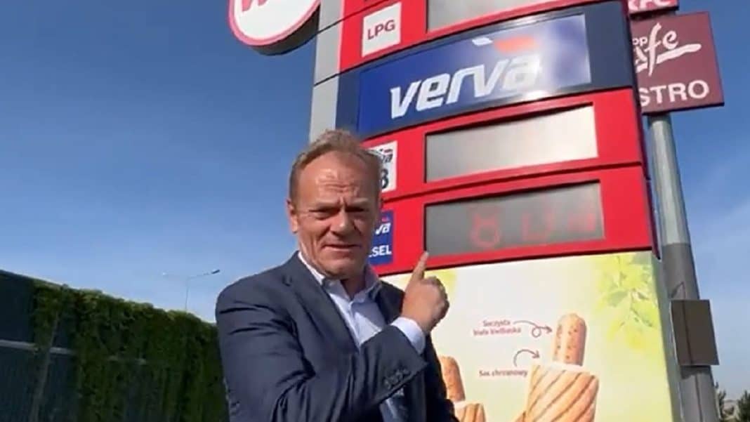 Mężczyzna wskazuje na wyświetlacz cen paliw na stacji benzynowej, w tle reklama i niebo.