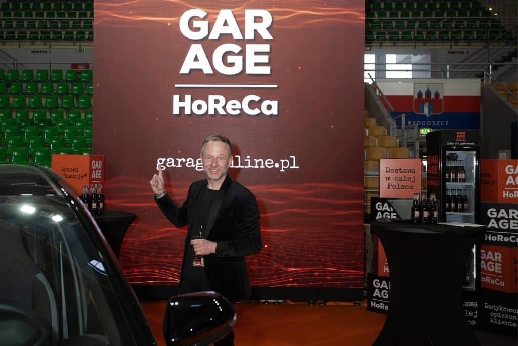Mężczyzna uśmiecha się i wskazuje na ekran z napisem 'GARAGE' podczas wydarzenia w hali sportowej.