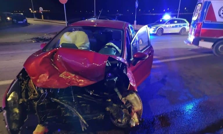 Rozbity czerwony samoch&oacute;d na skrzyżowaniu po zderzeniu, w tle służby ratunkowe z włączonymi światłami.