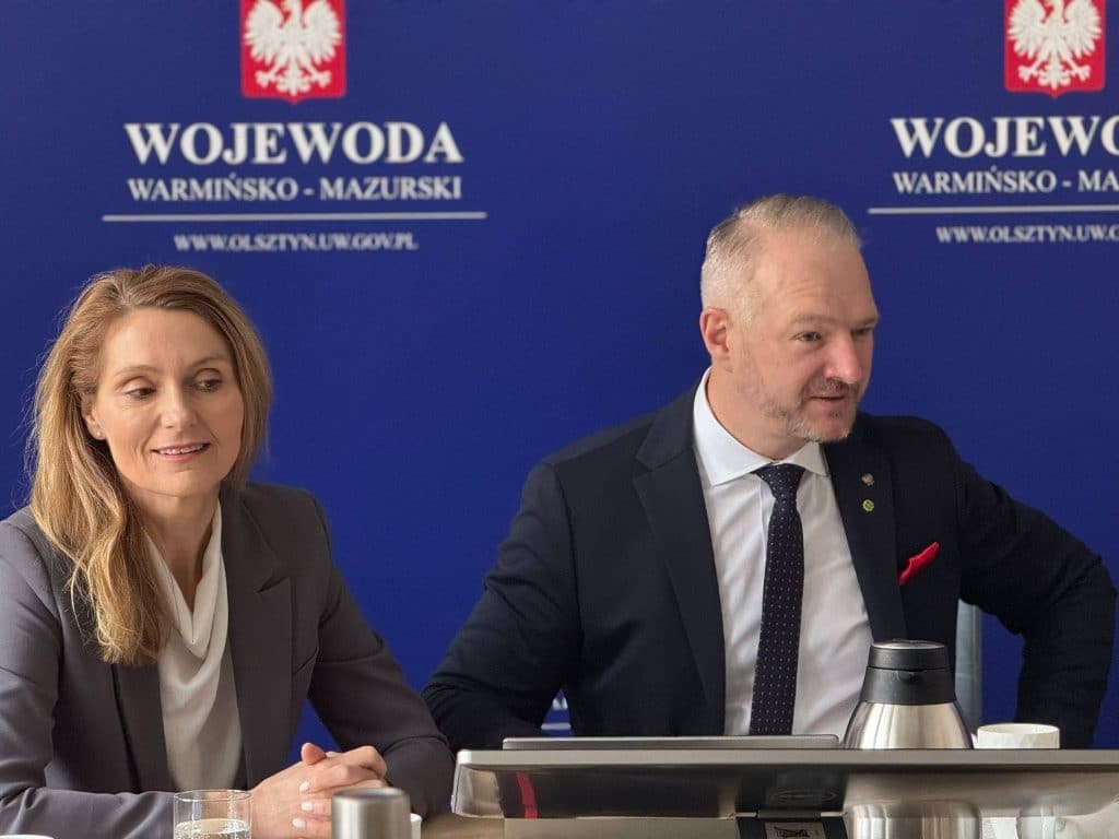 Dwoje os&oacute;b siedzi przy stole podczas konferencji, za nimi plakat z napisem 'Wojewoda Warmińsko-Mazurski'.