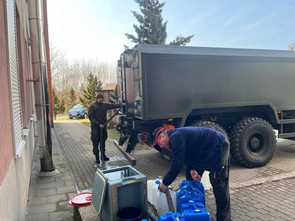 Wojsko i służby rozprowadzają wodę w regionie po skażeniu wody, w tle pojazd wojskowy i pracownicy.