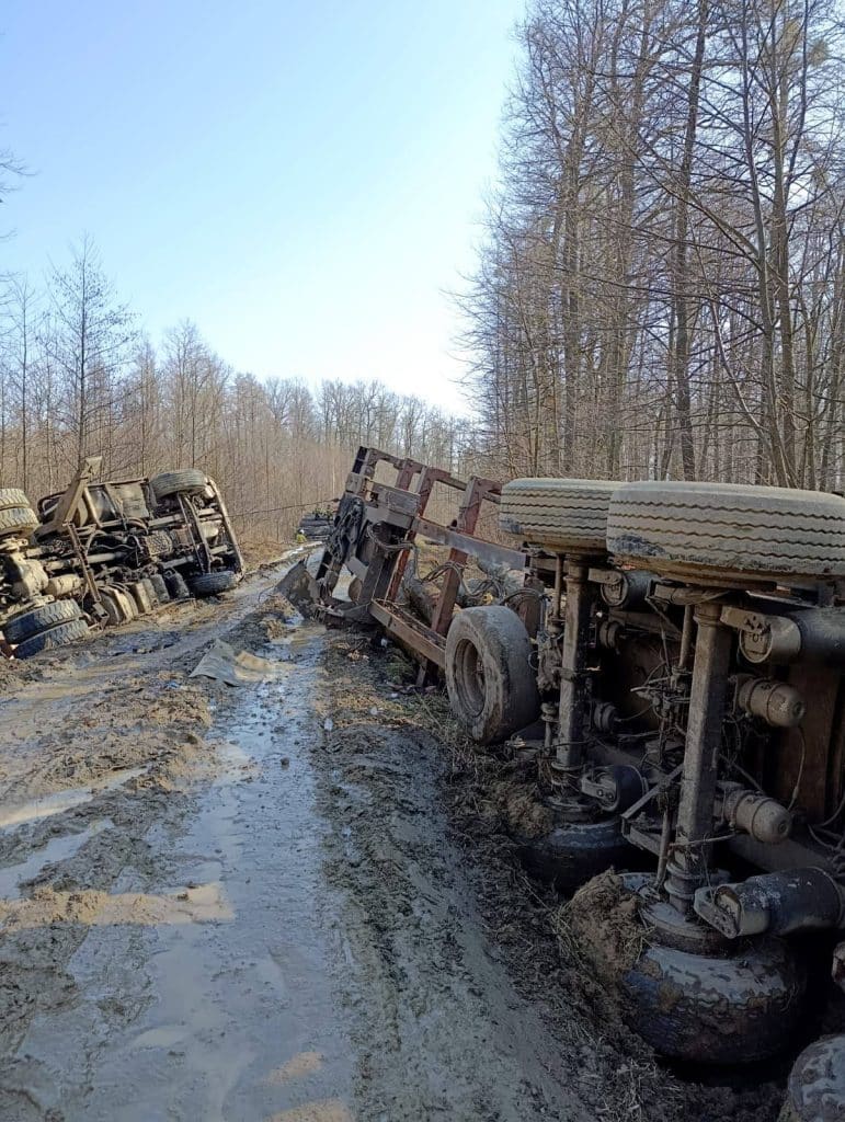 Rozbity pojazd ciężarowy leży na boku na leśnej drodze, wok&oacute;ł błoto i uszkodzenia.
