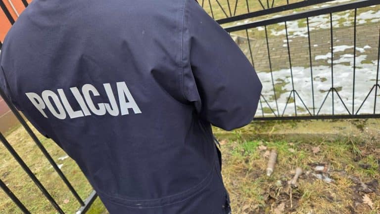 Policjant stoi przy ogrodzeniu szkoły podstawowej w Ełku, w tle śnieg i przedmioty na ziemi.