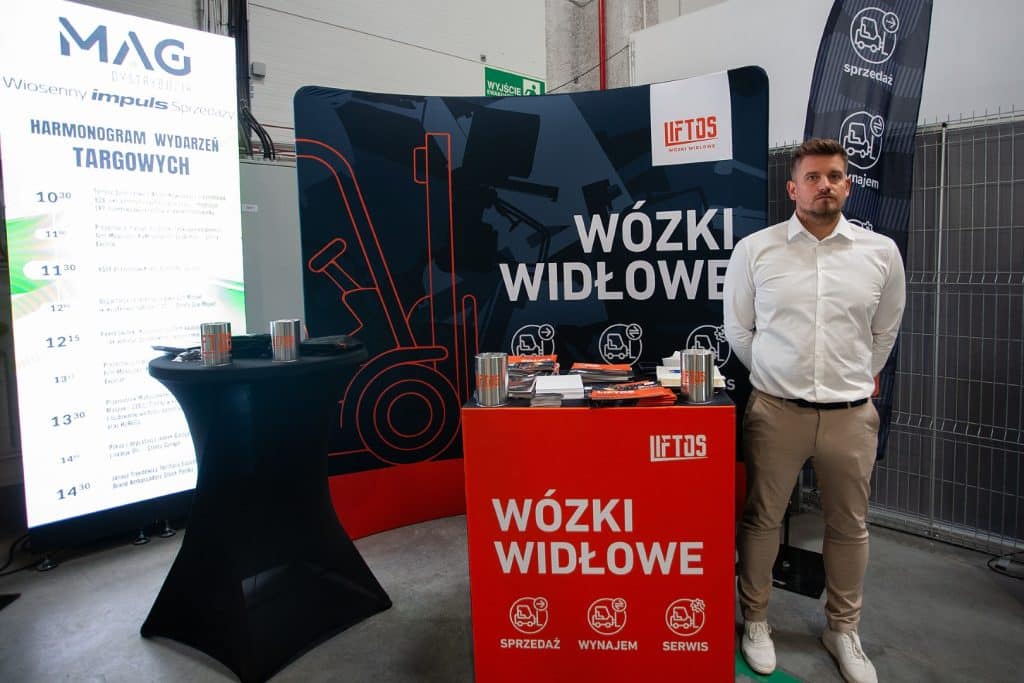 Stojący mężczyzna przy stoisku z w&oacute;zkami widłowymi na targach w Gdańsku, obok plansza z ofertą.