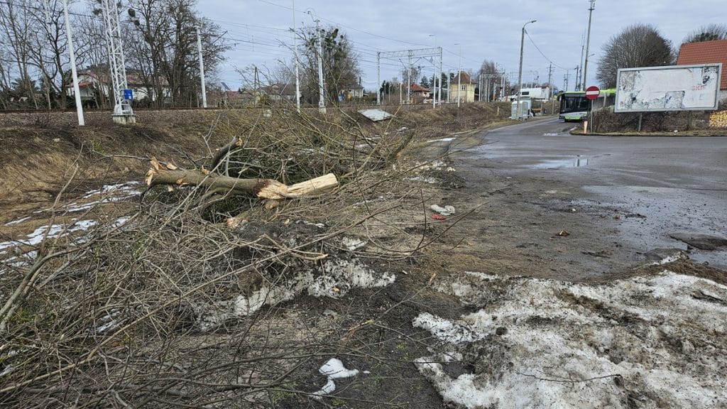 Zniszczony odcinek drogi z gałęziami i śniegiem, w tle widoczny autobus i tory kolejowe.