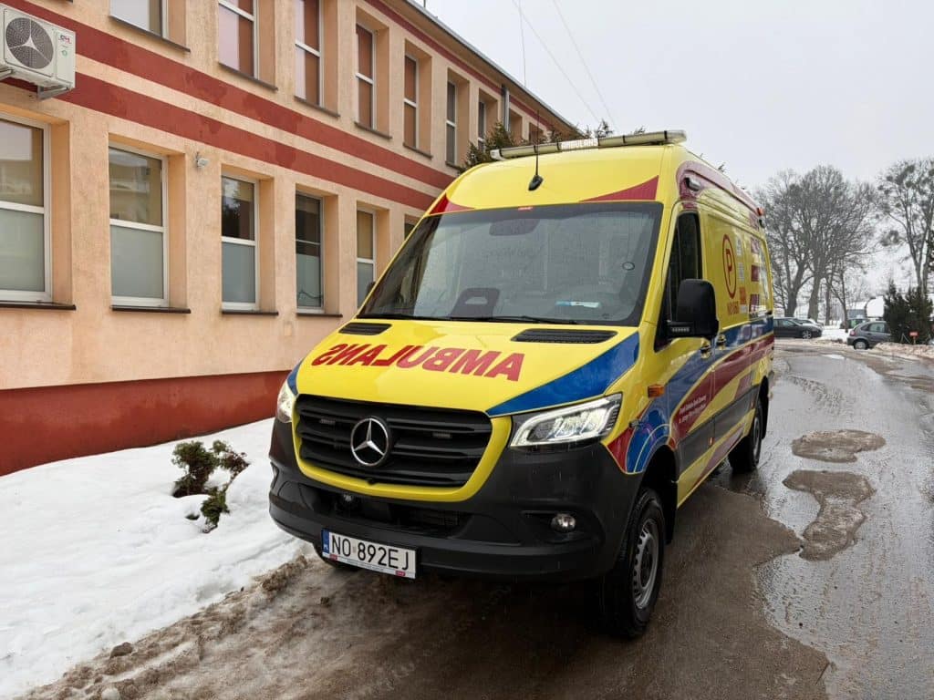 Ż&oacute;łty ambulans zaparkowany przed budynkiem szpitala w Dobrym Mieście, zimowa pogoda, śnieg i błoto na drodze.