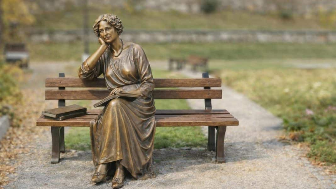 Pomnikowa rzeźba kobiety siedzącej na ławce w parku, z książkami obok.