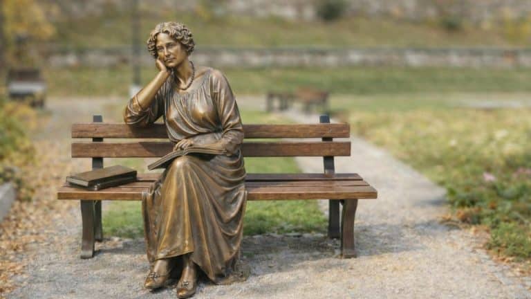 Pomnikowa rzeźba kobiety siedzącej na ławce w parku, z książkami obok.
