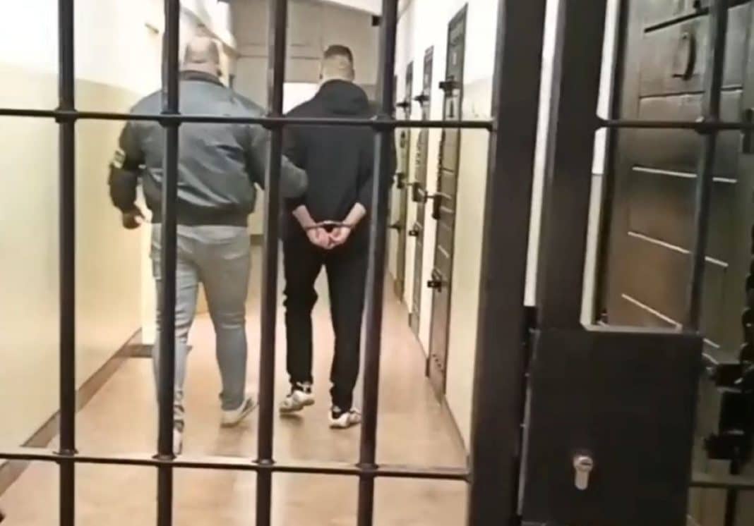 Mężczyzna w rękach policji, widoczny z tyłu, za kratami, podczas zatrzymania.