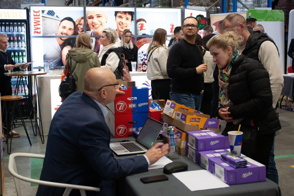 Uczestnicy targ&oacute;w biznesowych w Gdańsku rozmawiają przy stoisku z produktami spożywczymi i napojami.