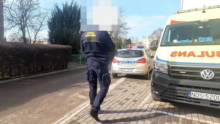 Miejski strażnik w uniformie stoi na chodniku obok radiowozu i ambulansu, na tle drzew i budynk&oacute;w.