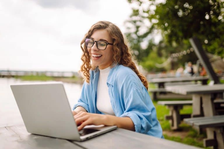 Kobieta pracująca na laptopie na świeżym powietrzu, w otoczeniu zieleni i ław, uśmiechnięta.