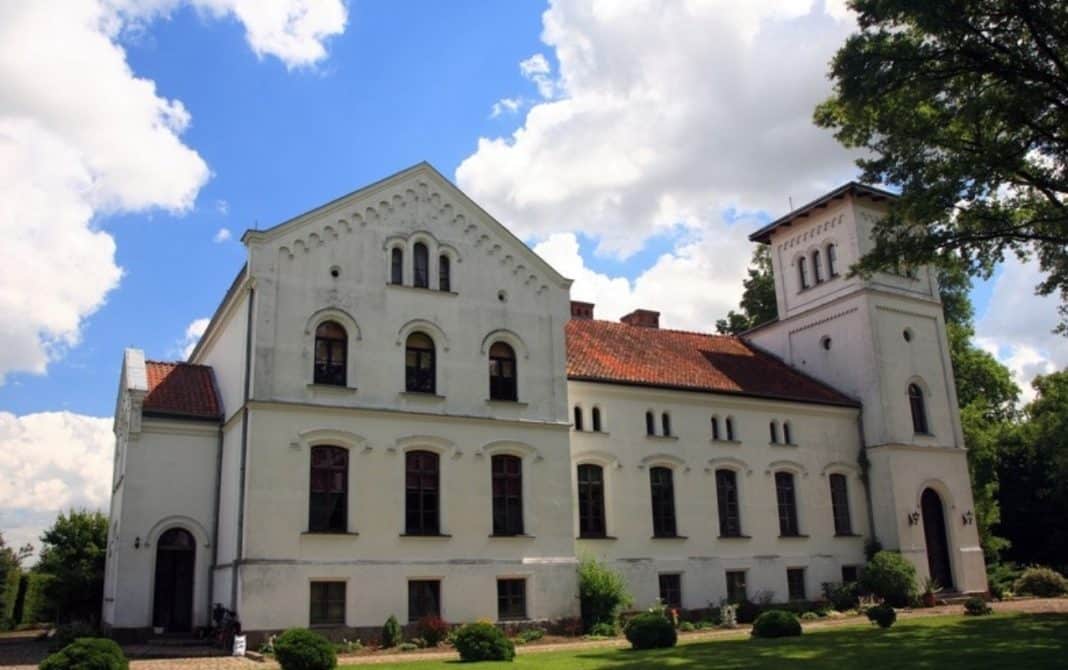 Zabytkowy pałac z XIX wieku na Warmii i Mazurach, biały, z wieżami, otoczony zielenią, pod chmurami.