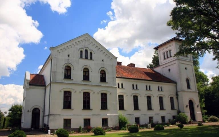 Zabytkowy pałac z XIX wieku na Warmii i Mazurach, biały, z wieżami, otoczony zielenią, pod chmurami.