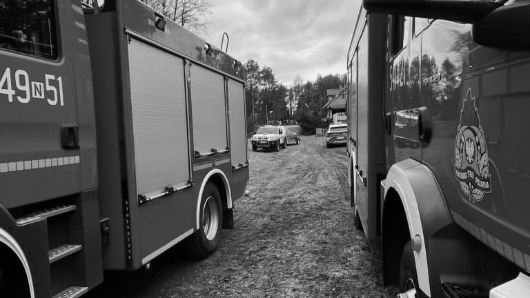 Dwa wozy strażackie na tle zamarzniętego jeziora i samochod&oacute;w, w tle drzewa i domy, pochmurny dzień.