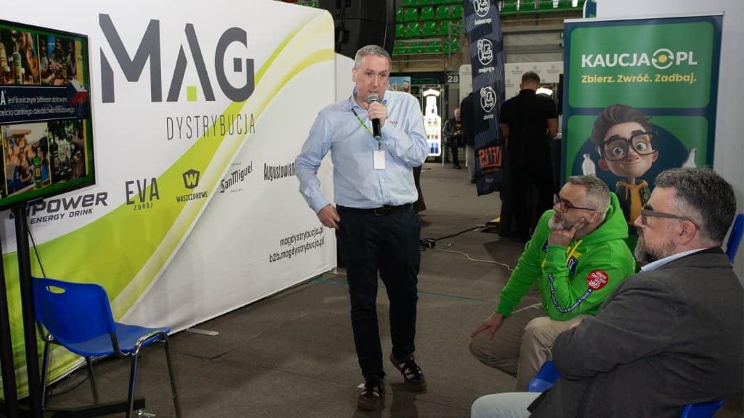 Mężczyzna z mikrofonem przemawia na targach branżowych w hali Urania, wok&oacute;ł stoją osoby słuchające.