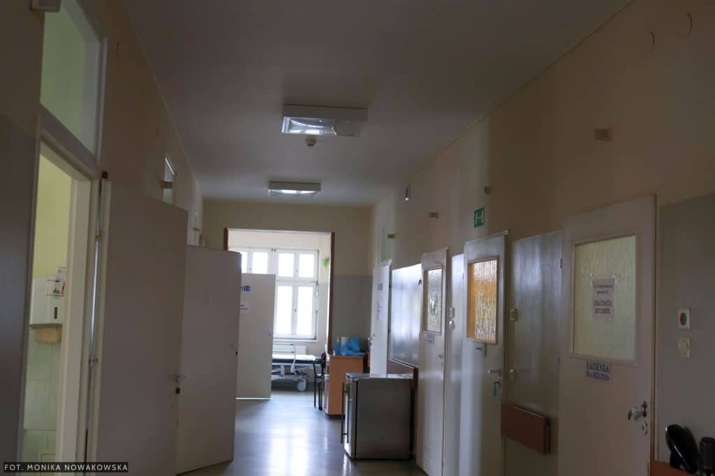 Widok korytarza w szpitalu z zamkniętymi drzwiami do sal i oknami na końcu.