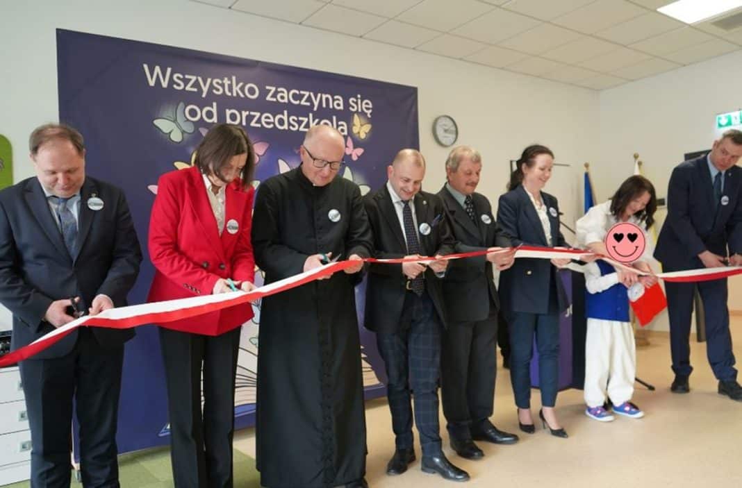 Grupa os&oacute;b podczas oficjalnego otwarcia nowego przedszkola w Jonkowie, z czerwonym tańczącym wstążką.