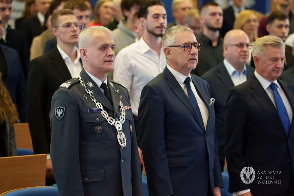Uczestnicy uroczystości na sali, w tym wojskowy w mundurze i dwaj mężczyźni w garniturach, podczas inauguracji nowego kierunku studi&oacute;w.