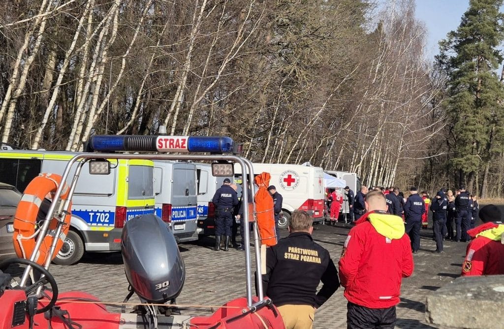 Grupa służb ratowniczych i policji na miejscu akcji poszukiwawczej w lesie, w tle drzewa i pojazdy ratunkowe.