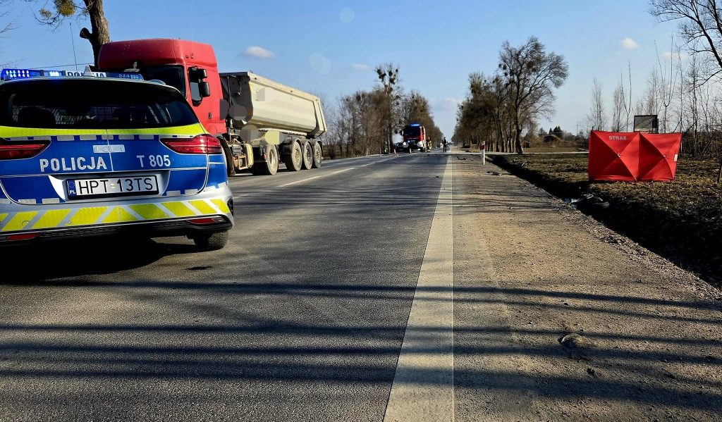 Widok na miejsce wypadku na drodze krajowej z radiowozem i ciężar&oacute;wką, w tle służby ratunkowe.