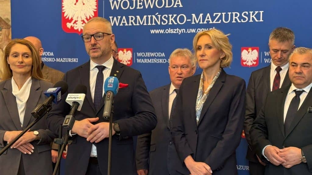 Grupa urzędnik&oacute;w podczas konferencji w wojew&oacute;dztwie warmińsko-mazurskim, przed tablicą z logo wojewody.