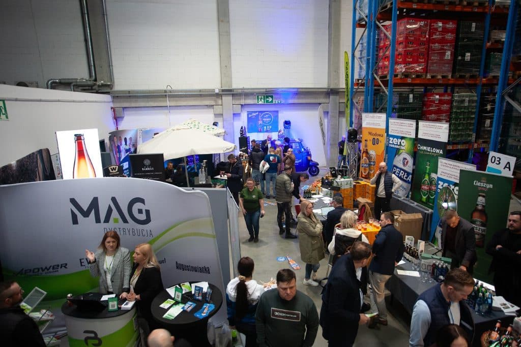 Tłum uczestnik&oacute;w na targach biznesowych w hali magazynowej, stoiska z produktami i rozmowy biznesowe.