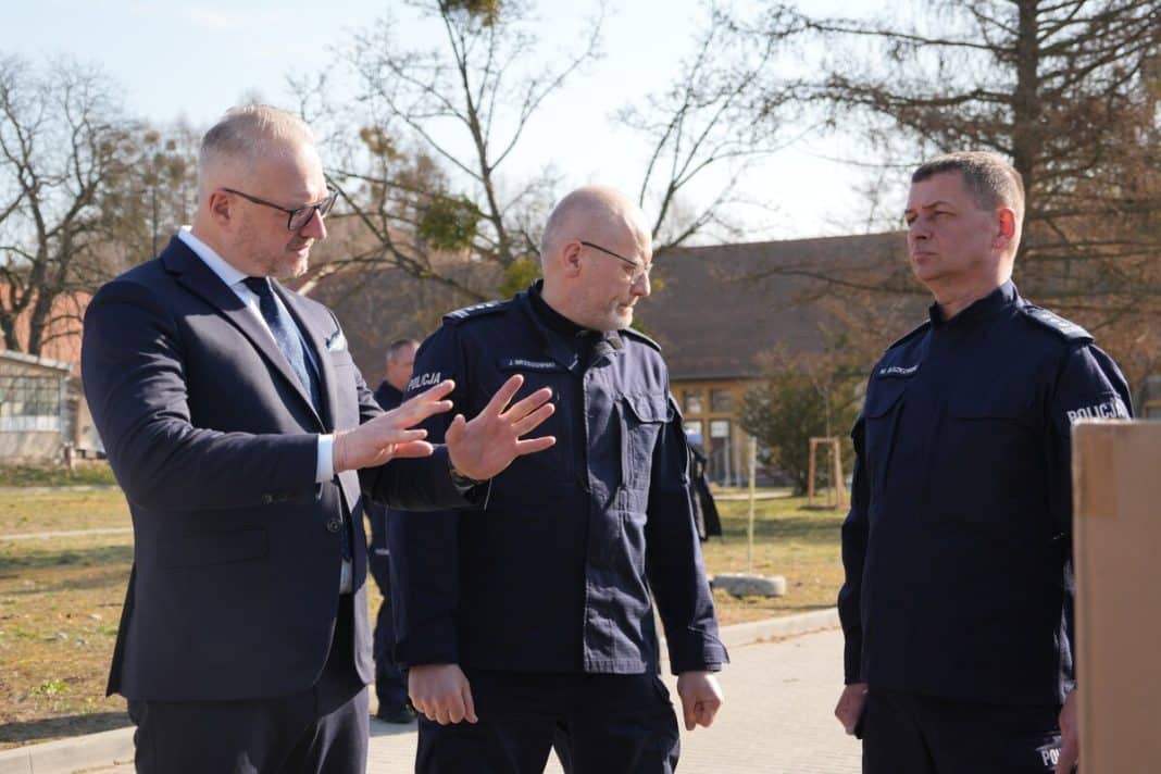 Policjanci w mundurach rozmawiają na zewnątrz, w tle drzewa i budynki, w słoneczny dzień.
