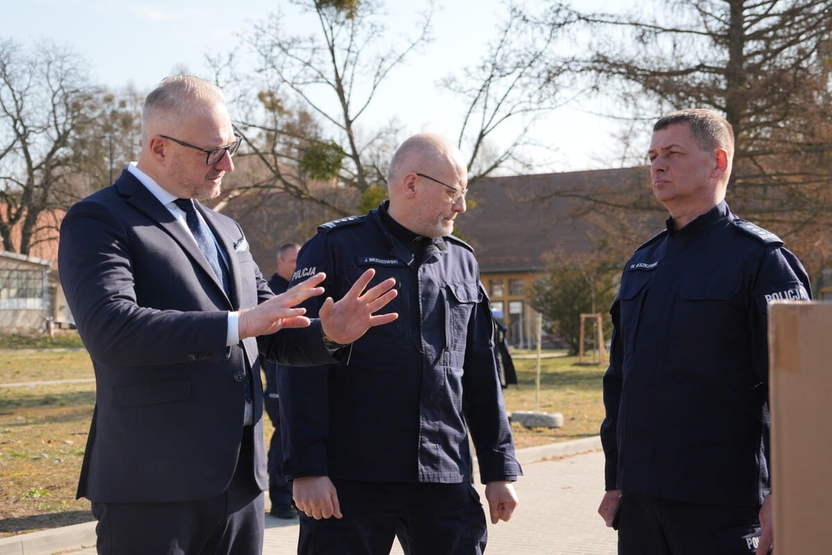 Policjanci w mundurach rozmawiają na zewnątrz, w tle drzewa i budynki, w słoneczny dzień.