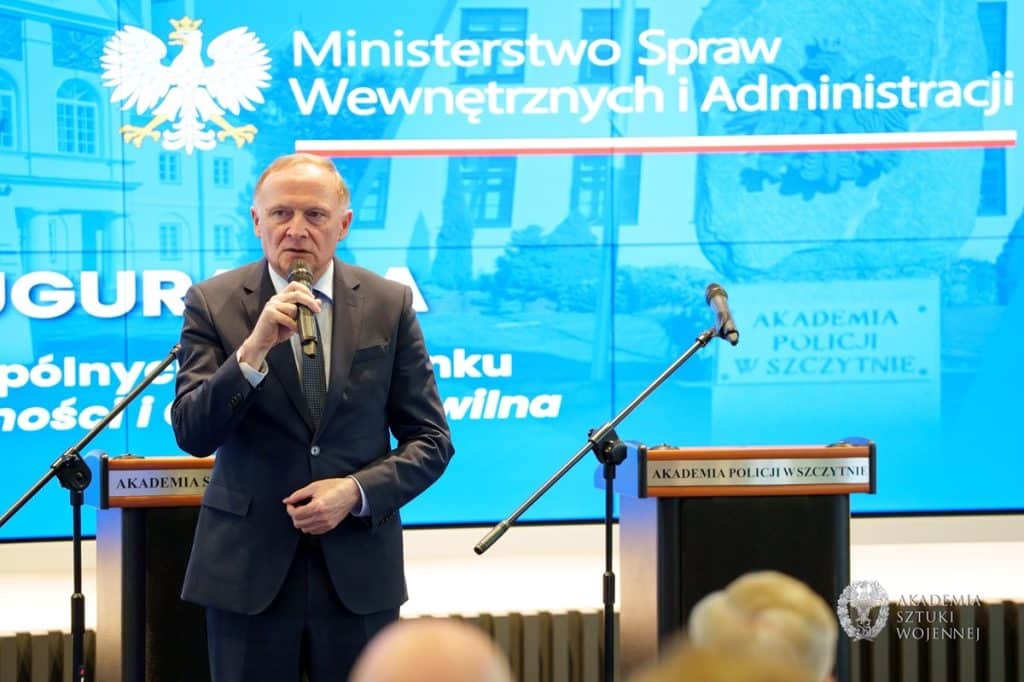 Mężczyzna w garniturze przemawia do publiczności podczas wydarzenia w Szczytnie, za nim ekran z napisem o nowym kierunku studi&oacute;w.