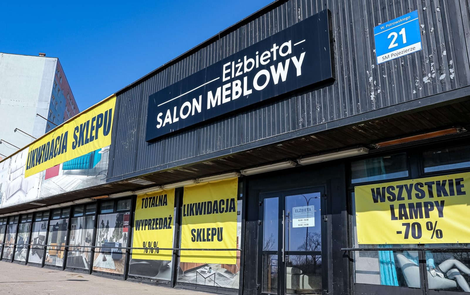 Widok na zewnątrz salonu meblowego Elżbieta z dużymi plakatami wyprzedażowymi na oknach.