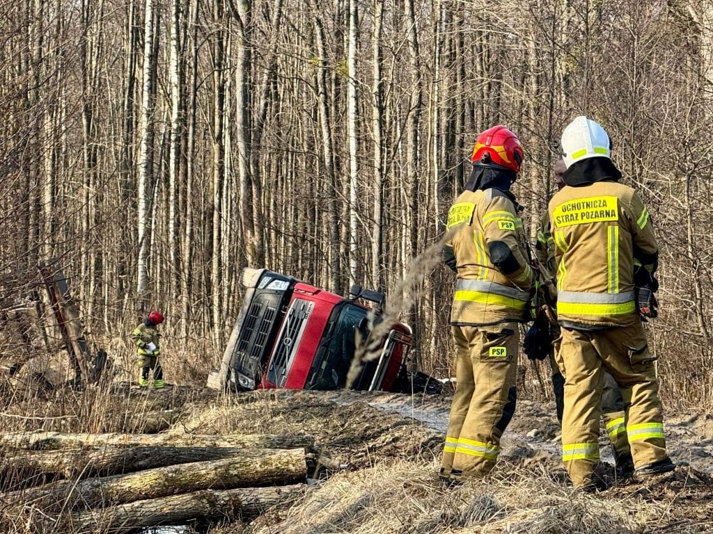 Strażacy na leśnej drodze obok przewr&oacute;conej ciężar&oacute;wki w rowie.