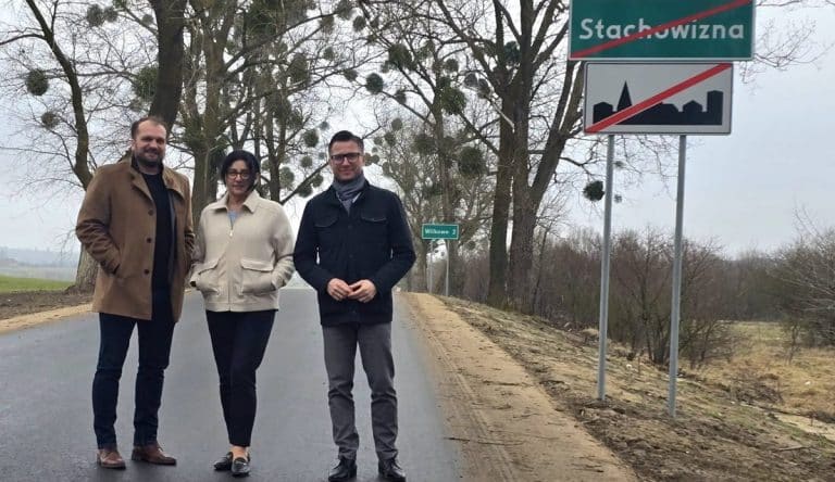 Trzech ludzi stoi na nowo wyremontowanej drodze w pobliżu znaku Wilkowo, w tle drzewa i tablica Stachowizna.