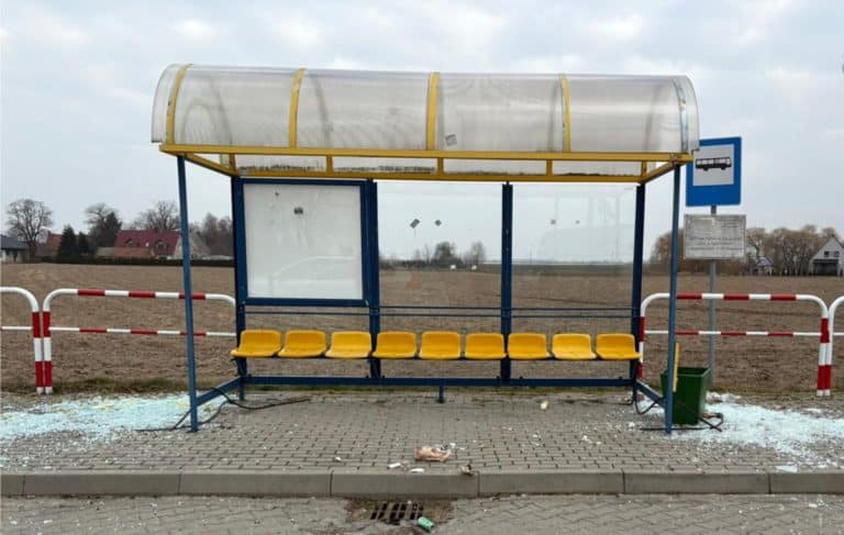 Zniszczona wiata przystankowa z rozbitym szkłem i odłamkami na chodniku, w tle puste pole i domy.