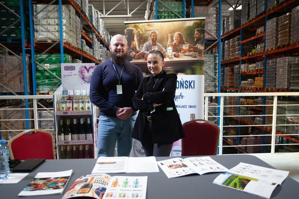 Uczestnicy targ&oacute;w w hali magazynowej, stoją przy stoisku z produktami i materiałami promocyjnymi.