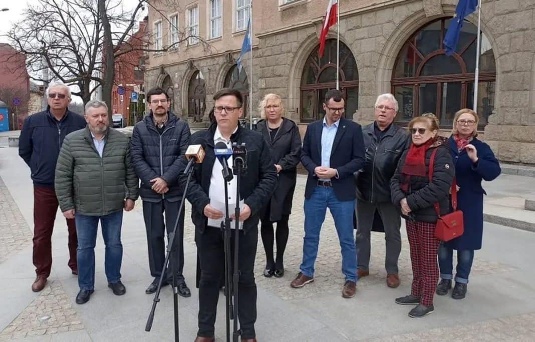 Grupa ludzi stojących na ulicy podczas konferencji prasowej przed budynkiem z flagami.