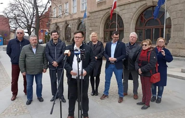 Grupa ludzi stojących na ulicy podczas konferencji prasowej przed budynkiem z flagami.