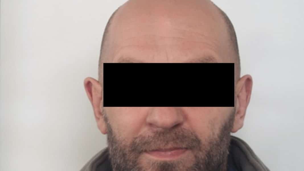 Zdjęcie poszukiwanego mężczyzny z zasłoniętymi oczami, kr&oacute;tkie włosy i broda, na tle białej ściany.