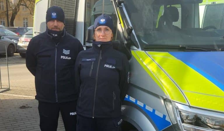 Dwoje policjant&oacute;w stoi obok radiowozu na ulicy w Olsztynie, w tle zaparkowane samochody.