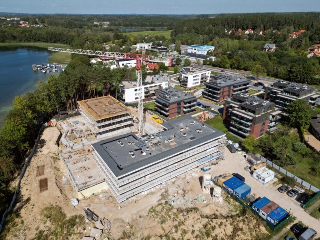 Budowa nowych budynk&oacute;w nad jeziorem Ukiel, w tle las i zabudowa mieszkaniowa.