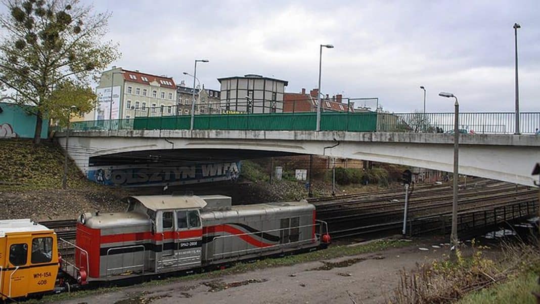 Wiadukt nad torami w Olsztynie, widok z dołu, z pociągiem na torach i otaczającą infrastrukturą.